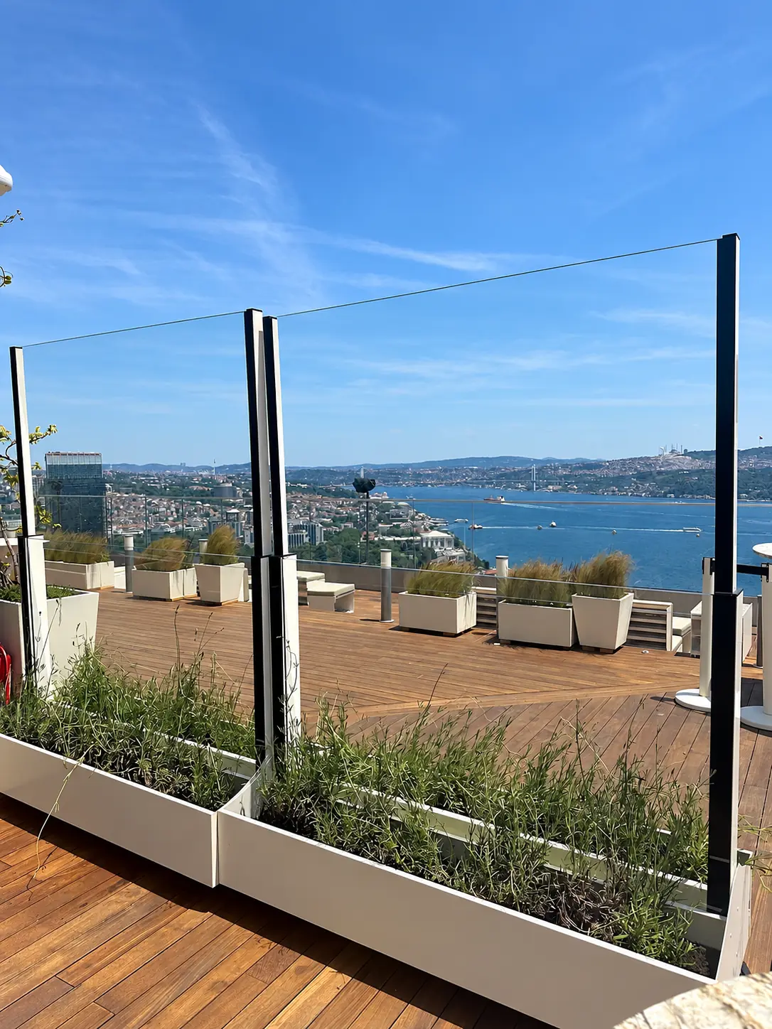 Două module de paravan antivânt din sticlă, cu panourile superioare ridicate și jardiniere albe, pe o terasă cu vedere la mare.