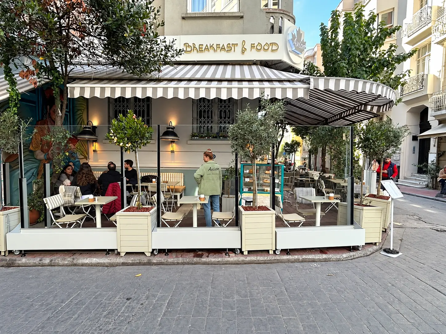 Module de paravan antivânt din sticlă cu baze albe în fața unei terase de restaurant, între jardiniere cu arbori.