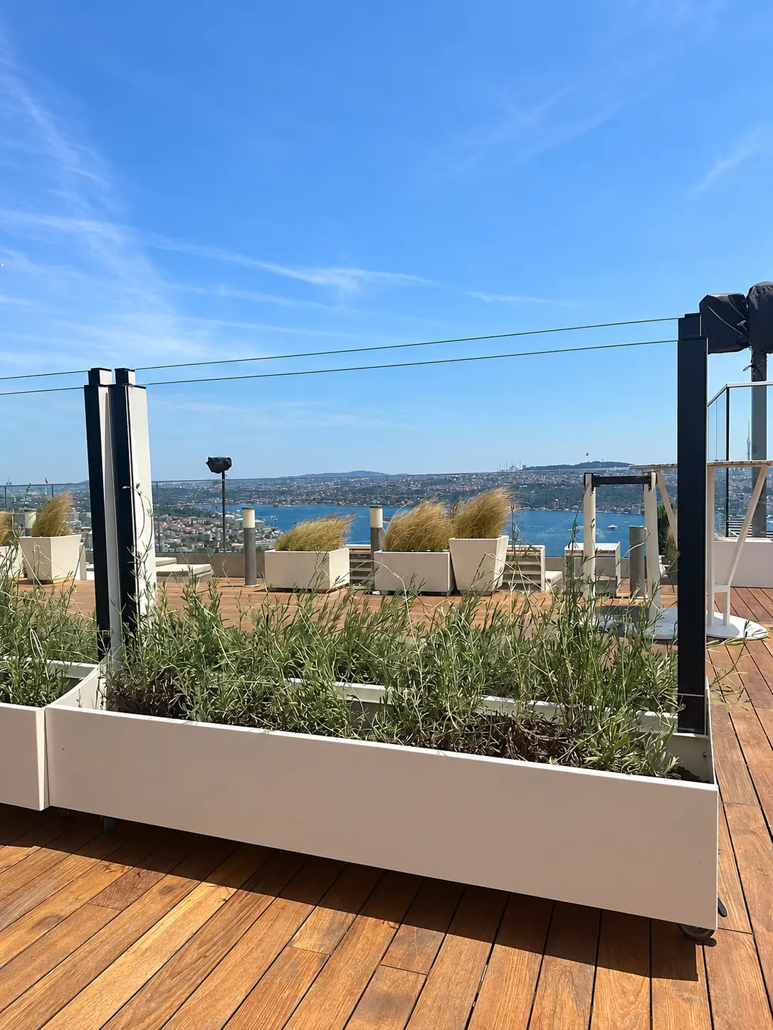 Paravan antivânt din sticlă cu panou superior într-o poziție mai joasă, deasupra unei jardiniere albe cu plante, pe o terasă cu vedere la mare.